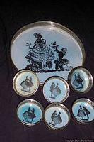 Oval light blue platter with black silhouette of two people in English Royalty style dress, surrounded by a silver metal rim. Six round light blue coasters with various black silhouette figures matching platter design.