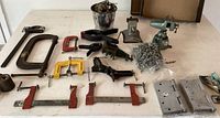 Photo showing various clamps, green small bench vise, metal chain, spray nozzle, and miscellaneous hardware items spread out on table.