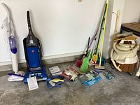 Full view of lot including Hoover upright vacuum, Max floor polisher, shop vac, stick vacuum, brooms, dusters, brushes, dustpans, and cleaning supplies.