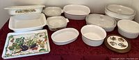 All Corning Ware baking dishes and serving tray arranged on red tablecloth: rectangular, oval, round shapes, some with Harvest pattern, some plain white, glass and ceramic lids, and a wooden-trimmed ceramic trivet visible.
