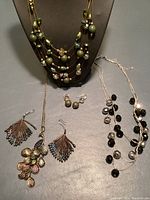 View of several necklaces including multi-strand AB crystal necklace with green beads, two thin wire sparkly bead necklaces, broken necklace chain, and pairs of earrings including vintage peacock feather clips and green bead clips arranged on a gray surface and necklace display stand.