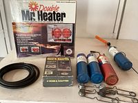 Photo displaying Mr. Heater Double Radiant Propane Heater original box, 4' hose and adapter in packaging, coiled black hose, 4 propane tanks (3 blue, 1 red), an orange-handled weed burner and several metal clamps and chains.