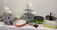 Wide view showing entire lot including Pyrex dishes, tiered serving trays, fondue pot, condiment server, and skewers on white fabric surface