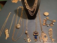 Overview of various gold-tone costume jewelry pieces including layered chain necklace on display stand, several long chain necklaces with pendants, charm bracelets and compact on a flat surface.