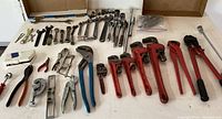 Overview of pipe wrenches, adjustable wrenches, pliers, and socket wrenches laid out on table for lot examination.