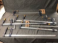 Full view of six large metal bar clamps arranged on a dark fabric background, showing variations in handle styles and bar lengths.