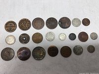 Full overview of 21 older coins and tokens laid out on a white cloth, various sizes and colors including bronze and silver toned items. Some have visible perforations or holes.