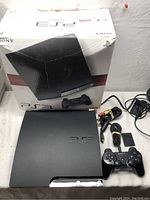 Sony PlayStation 3 console on table next to controller, cables, and original box. Console front and surface visible.