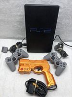 Front view of PlayStation 2 console with three controllers, orange light gun, and power adapter cable.