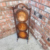 Three-tier wooden folding cake stand in folded position leaning against brick wall outdoors. Shows top handle, turned spindles, and circular trays with surface patina and wear.