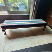 Full side view of the antique kneeling bench showing the upholstered top with striped fabric, wooden frame, and cabriole legs on tile floor next to window.