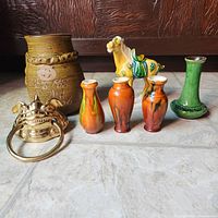Full view of the collection showing seven items on tile floor: large brown vase with embossed characters, three small ochre-brown vases, green vase, camel figurine, and brass ring.