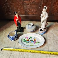 Overview of the lot items on tile floor with measuring tape showing approximate sizes: male geisha figurine in red and black standing, white Guanyin figurine seated on fish base, small blue and white porcelain vase, small gray round vase, hand painted plate.