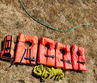 Four orange adult lifejackets, one small pet lifejacket, and yellow rope laid out on grass.