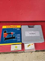 Samona stapler in original box with Bynford stapler gray carrying case and packs of staples and nails in front of red bench.