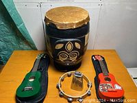 Photo showing vintage carved and painted drum, green ukulele, red ukulele with sticker, tambourine, and cowbell all placed on a table
