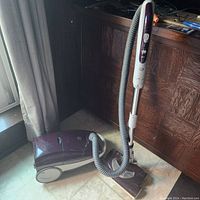 Photo of Kenmore canister vacuum with attached hose, wand, and floor brush on tiled floor near window and wooden furniture.