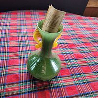 Green Murano glass vase with bulbous base and slender neck, showing yellow curled handles and certificate inside. Positioned on red plaid tablecloth, photographed in natural light from multiple angles to capture detail and visible chip on one handle.