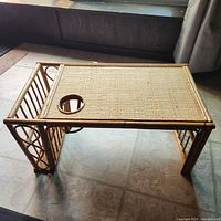Angled view of rectangular breakfast tray/lap table showing woven reed surface with circular cup holder cutout and bamboo frame structure with supporting legs.