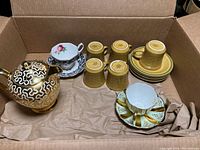 Tea time lot showing various cups, saucers, and a teapot inside a cardboard box with brown packing paper