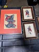 Three framed cross stitch artworks laid on a flat surface, showing the Scotty dog on the left in an orange frame and the two Highland dancers on the right in ornate gold frames.