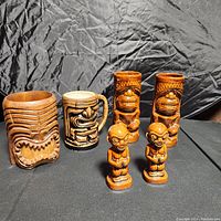 Six vintage tiki themed items arranged on table with black backdrop: three tiki mugs including one with handle, two small tiki figurines, and two tall tiki mugs.