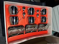 Photo of the full set displayed in its box showing 6 cups, 6 saucers, and 6 spoons on orange insert.