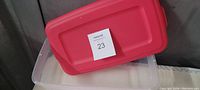 Clear Sterilite rectangular plastic container with red lid and auction sticker labeled 23 on lid.