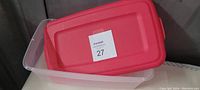 Clear plastic rectangular storage container with a bright red lid placed on top at an angle, showing the label 27 on the lid indicating lot number.