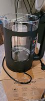 Electric kettle with glass body inside black plastic frame with handle and cord on cardboard box