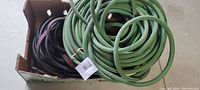 Two coiled garden hoses, green and black with red stripe, inside a cardboard box on concrete floor.