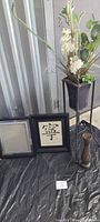 Framed 'Serenity' picture and empty frame side by side with vase and faux plant arrangement on metal stand nearby.