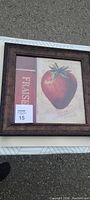 Framed square wall artwork with a vintage strawberry illustration and French text, photographed outdoors on gray pavement.