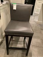 View of one gray leather bar stool showing padded seat and backrest, dark wood square legs, and silver nailhead trim at seat edge.