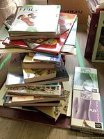 Top view showing a tall stack of books and Pilates DVDs on a wooden floor with a few boxes in background.