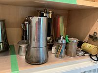 Shelf view showing Italian espresso coffee maker pot, metal coffee mugs with golden accents, container with multicolored plastic straws, and metal shaker.
