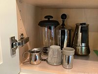 Photo of kitchen cabinet shelf with large and small French presses, metal creamer pitcher, metal filter, and small stainless steel container, showing item variety and condition.