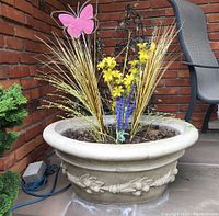 Full view of the concrete planter showing faux plants, soil, and pink butterfly stake, placed against a brick wall with a chair in background.