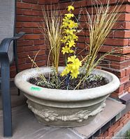 Front angled view of large round concrete planter with faux yellow flowers and grasses inside. Planter has decorative raised floral motifs around exterior.