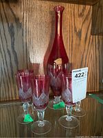 Photo of cranberry glass decanter with six matching stemmed glasses on wooden shelf.