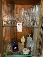 Wide view of shelf containing assorted crystal stemware, crystal pitchers, ceramic plate, alabaster compote, luster trinket box, small decorated glass vase, and Swarovski figurine on blue box.