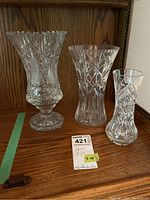 Three crystal vases on wooden surface showing front and side views, ranging 7 to 10 inches tall with visible intricate cut patterns.