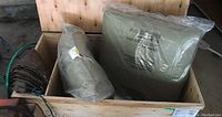 Photo showing a wooden box containing a folded dark green tarp and two cushions wrapped in clear plastic.