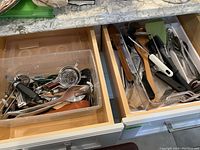 First drawer containing assorted kitchen utensils including peelers, strainers, forks, and spatulas in a plastic bin and organized compartments.