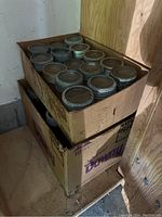 Two cardboard boxes stacked, each filled with Mason jars with metal lids. Jars vary in size, surface clean with some dust and slight signs of storage.