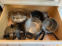 Photo of various stainless steel and metal cookware including stock pot, saucepans, mixing bowl, steamer insert, and lids in a drawer.