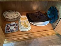Shelf with assorted serving platters, trays, and plates including floral rimmed round plates, white rectangular trays with wheat motif, dark textured square platters, and large blue-green sculpted ceramic plate with gold trim.
