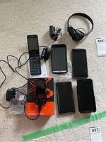 Photo of six cell phones displayed on carpet, including flip and touchscreen models with accessories such as wired headphones and chargers.