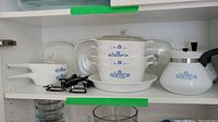 Shelf display of Corning Blue Cornflower pattern cookware and bakeware including saucepans, baking dishes, pile of stacked glass dishes, kettle, and lids.