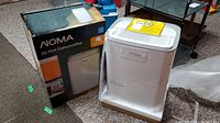 NOMA dehumidifier unit with original box visible behind, the unit retains protective foam and shrink wrapping, appears new.